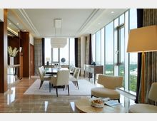 Elegantes Wohnzimmer mit modernem Essbereich, großen Fensterfronten, die eine Aussicht auf eine Stadt und Grünflächen bieten. Helle Möbel, ein Teppich unter dem Esstisch sowie Holzelemente bestimmen das Design, und eine Stehlampe sowie Dekorationen sorgen für Gemütlichkeit.