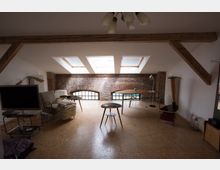 Dachgeschoss-Wohnraum mit sichtbaren Holzbalken, einer Backsteinwand mit abgerundeten Fenstern und drei Dachfenstern, die den Raum erhellen. Der Raum ist mit Möbeln wie einem Sofa, Tischen, einem Fernseher und einem Schreibtisch eingerichtet und hat einen hellen Holzboden.