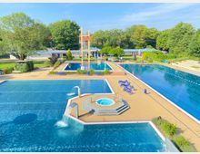 Ein Freibad mit mehreren Becken, darunter ein großes Schwimmerbecken und ein kleineres Becken mit Sprudeldüsen. Im Hintergrund sind eine Liegewiese, Bäume und ein flaches Gebäude zu sehen, die Umgebung ist von Grünflächen und Sitzbereichen umgeben.
