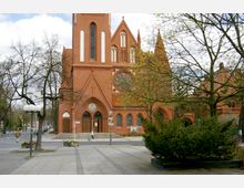 Backstein-Kirche im neugotischen Stil mit spitzem Kirchturm, großen Spitzbogenfenstern und einem runden Rosenfenster über dem Eingangsportal. Im Vordergrund sind ein gepflasterter Platz, Bäume mit frischem Laub und eine Straßenkreuzung zu sehen.