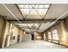 Industriehalle mit unverputzten Backsteinwänden und großen Fenstern, durch die Tageslicht einfällt. Der Raum verfügt über eine hohe Decke mit sichtbaren Metallträgern, mehreren Dachfenstern und einer zentral aufgehängten runden Uhr.