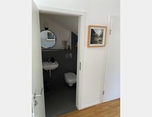 Ein kleiner, modern gestalteter Toilettenraum mit grauen Fliesen, einem Wandwaschbecken und einem Wand-WC. An der weißen Wand außerhalb des Raumes hängt ein gerahmtes Foto, der Boden besteht aus hellem Holzparkett.
