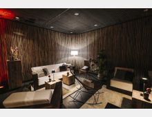 Ein gemütlicher Loungebereich mit moderner Einrichtung, darunter weiße Sofas, Holztische und ein dekorativer Sessel. Die Wände sind mit dunklem Holz vertäfelt, beleuchtet durch eine Stehlampe; Zimmerpflanzen und kleine Dekorationen ergänzen die Atmosphäre.