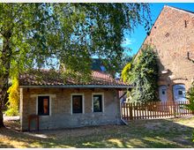 Kleines Gebäude aus Backstein mit roten Dachziegeln, drei Fenstern und einem einzelnen Holztisch daneben. Im Hintergrund ein größeres Backsteingebäude mit bogenförmigen Türen und einem bewachsenen Zaun, umgeben von Bäumen und Rasen.