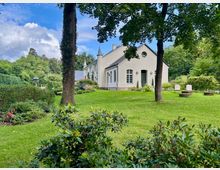 Helles, villaähnliches Gebäude mit Satteldach, Rundfenster und kleinen Türmchen, umgeben von einem gepflegten Garten mit Rasenflächen, Sträuchern und großen Bäumen. Im Vordergrund liegen Beete und Hecken, rechts stehen zwei schlichte Sitzgelegenheiten auf der Wiese.
