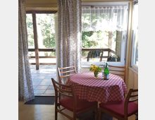 Essbereich mit einem runden Tisch, bedeckt mit einer rot-weiß karierten Tischdecke, umgeben von vier Holzstühlen mit roter Polsterung. Im Hintergrund ist durch eine verglaste Terrassentür und ein Fenster eine überdachte Terrasse mit Fliesenboden und grünem Außenbereich zu sehen.