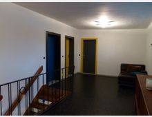 Flur mit Treppenaufgang aus Holz und einem schwarzen Geländer, das mit farbigen Kreisen verziert ist. Drei Türen mit unterschiedlich farbigen Rahmen (gelb und blau) sowie eine braune Ledercouch und ein Sideboard prägen den Raum.