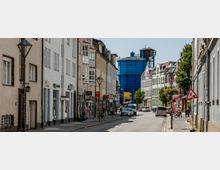 Straßenansicht in einer Stadt mit historischen, mehrstöckigen Häusern auf beiden Seiten, verschiedenen Geschäften im Erdgeschoss und einem großen blauen Gasometer im Hintergrund. Autos parken am Straßenrand, während Fußgänger auf den Gehwegen unterwegs sind; vereinzelt stehen Laternen und blühende Blumenampeln.