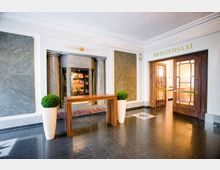 Heller Eingangsbereich mit schwarzem, glänzendem Boden, weißen Wänden und zwei dekorativen grauen Marmortafeln. Rechts befindet sich eine Tür aus Holz und Glas mit der Aufschrift „Meistersaal“ darüber, während in der Mitte ein schmaler Holztisch zwischen zwei großen weißen Pflanzgefäßen steht.