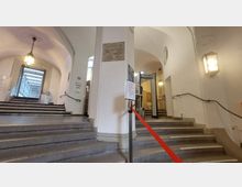 Treppenhaus mit hellen, gewölbten Decken und Wandleuchten. In der Mitte trennt ein rotes Absperrband den Zugang zu einem Metallscanner; seitlich führen Treppen mit Handläufen in verschiedene Richtungen.