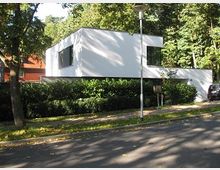 Moderner, kubischer Bau mit weißer Fassade, großen Fensterfronten und flachem Dach, umgeben von Bäumen und Büschen in einer grünen Wohngegend. Im Vordergrund führt eine asphaltierte Straße vorbei.
