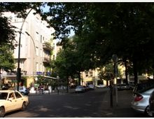 Straßenszene in einer Stadt mit Bäumen, die den Gehweg und die Straße beschatten. Mehrstöckige Wohnhäuser mit Balkonen und Geschäften im Erdgeschoss säumen die Straße; geparkte Autos und ein vorbeifahrendes Taxi sind sichtbar.