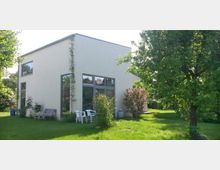 Modernes Wohnhaus mit Flachdach und cremefarbener Fassade. Das Gebäude verfügt über große Fenster und ist von einem Garten mit Rasen, Sträuchern und Bäumen umgeben.