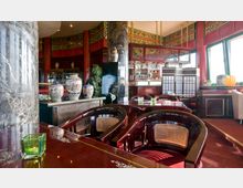 Innenraum eines asiatisch gestalteten Restaurants mit roten Holzelementen, dekorativen Vasen auf einem Holztresen, dunklen Stühlen mit geflochtener Lehne und detailreichen Verzierungen an den Wänden. Im Hintergrund befinden sich eine Bar, Trennwände im japanischen Stil und große Fenster mit grünen Rahmen.