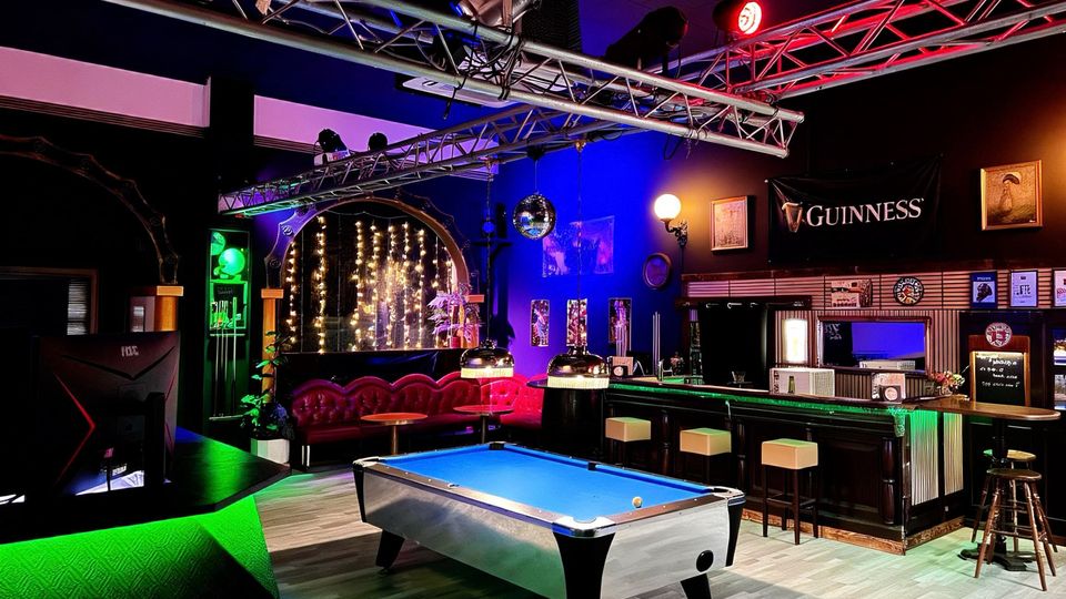 Innenraum einer Bar im Pub-Stil mit Billardtisch im Vordergrund, langer Theke mit Hockern und rotem Leder-Sitzbereich an der Rückwand. Über dem Raum hängen Metall-Traversen mit Bühnenleuchten; farbige Lichtakzente und Dekorationen wie Spiegelkugel und Guinness-Banner prägen die Atmosphäre.