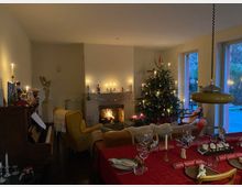 Gemütliches Wohnzimmer mit Kaminfeuer, dekoriert mit Kerzen und einem geschmückten Weihnachtsbaum neben großen Fenstern zur Terrasse. Im Vordergrund steht ein gedeckter Esstisch mit roter Tischdecke, Gläsern und Kerzen; links sind ein Klavier und Sitzmöbel zu sehen.