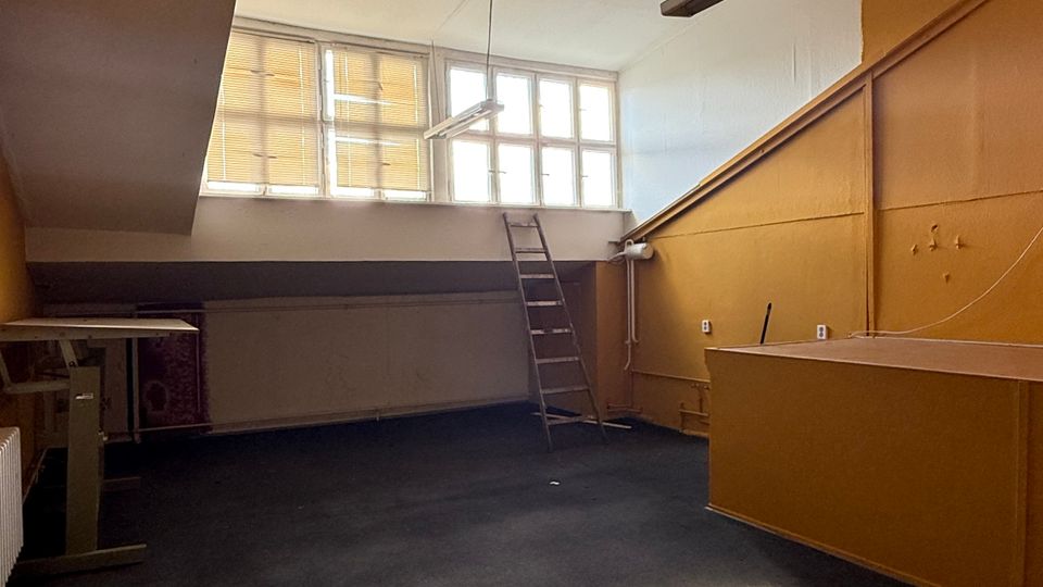 Leerer Innenraum mit schräger Decke, hohen Fenstern an der Stirnseite und hängenden Leuchtstofflampen. An der rechten Seite steht ein einfacher Tresen, daneben führt eine Leiter zu einer erhöhten Ablage unter den Fenstern.