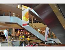 Innenansicht eines modernen Einkaufszentrums mit mehreren Rolltreppen, Bannern mit Blumendekor und Geschäften im Hintergrund. Sichtbar sind unter anderem Mode- und Sportgeschäfte sowie Werbeschilder.