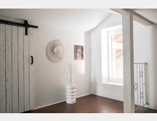 Minimalistisches Interieur mit weiß gestrichenen Wänden, einem Holzschiebetor auf der linken Seite, einem kleinen Fenster, einer dekorativen Wandverzierung und einem weißen Beistelltisch mit Glasvase. Der Boden ist aus dunklem Holz, und eine Metallgeländerstrebe befindet sich rechts neben dem Fenster.