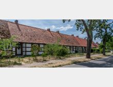 Langes Fachwerkhaus mit rotem Ziegeldach und mehreren Eingängen, umgeben von kleinen Vorgärten mit Zäunen und Bäumen, am Rand einer ruhigen Straße gelegen. Der Himmel ist leicht bewölkt, und die Vegetation wirkt sommerlich trocken.