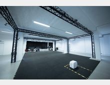 Ein großer, moderner Raum mit Metallstützstrukturen, schwarzem gepolstertem Boden und weißen Wänden. Im Hintergrund sind Schreibtische mit Monitoren und schwarzem Vorhang zu sehen, während ein mit Stoff bedeckter Würfel und gelbe Markierungen den Vordergrund betonen.