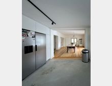 Offener Wohn- und Essbereich mit angrenzender Küche, weißer Einbauzeile und großem Edelstahl-Kühlschrank im Vordergrund. Im Hintergrund stehen ein langer Esstisch auf Holzboden, eine Pendelleuchte sowie eine Glastür mit Tageslicht; der Küchenbereich hat einen grauen Betonboden und schwarze Deckenschienenbeleuchtung.