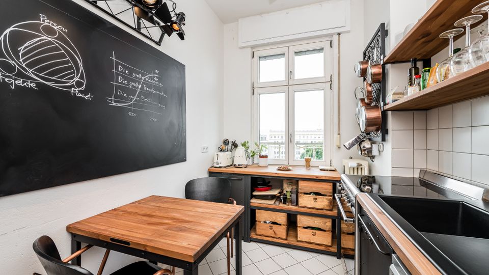 Kompakte, helle Wohnküche mit großem Fenster, Holzarbeitsflächen und einem kleinen Esstisch. Links hängt eine große Tafelwand mit Kreidezeichnungen, rechts sind offene Regale mit Gläsern, aufgehängten Töpfen sowie ein Herd und Spülbecken zu sehen.