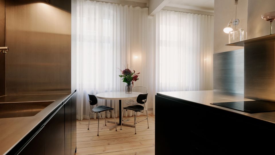 Moderne Küchen- und Essecke mit dunklen, minimalistischen Einbauschränken und einer großen Kücheninsel mit Kochfeld. Vor zwei großen Fenstern mit weißen, bodenlangen Vorhängen steht ein runder Tisch mit Stühlen auf hellem Holzboden.