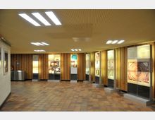 Ein hell beleuchteter Innenraum mit niedriger Decke, Holzwänden und gläsernen Ausstellungsvitrinen, die mit leuchtenden Informations- und Bildtafeln zu verschiedenen Themen bestückt sind. Der Boden besteht aus rötlich gemusterten Fliesen.