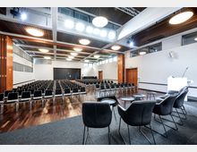 Großer Konferenzraum mit Reihen schwarzer Stühle auf einem Holzboden, einer Bühne mit schwarzen Sesseln und Tischen im Vordergrund sowie einer weißen Präsentationswand und Holzdetails an den Wänden. Die Decke ist mit Einbauleuchten und Holzpaneelen versehen.