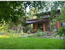 Holzhaus mit großen Fenstern, umgeben von einem üppigen Garten mit Rasen, Blumenbeeten und schattenspendenden Bäumen. Ein Sonnensegel ist über einer Terrasse gespannt, die zum Haus führt.