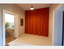 Ein Innenraum mit weißen Holzböden, einer orangefarbenen bodenlangen Vorhangwand und einer einzelnen Hängelampe an der Decke. Durch eine geöffnete Tür ist ein angrenzender Raum mit Holzboden, einem Tisch und bunten Stühlen sichtbar.