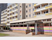 Ein mehrstöckiges Wohngebäude im Plattenbaustil mit gelben Fassadenelementen und Balkonen. Im Erdgeschoss befindet sich eine farbenfrohe Fensterfront mit horizontalen Streifen in Lila, Orange, Gelb und Grün; davor eine Treppe mit Geländer sowie bepflanzte Blumenkästen.