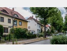 Straßenszene mit Reihenhäusern im traditionellen Stil, gekennzeichnet durch rote Ziegeldächer und helle Fassaden. Der gepflasterte Gehweg ist von Bäumen gesäumt, mit parkenden Autos und vereinzelten Fußgängern.