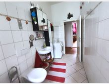 Narrow bathroom with white tiled walls and a diagonal-tile floor, featuring a sink with mirror and shelves, wall cabinets, and a toilet in the foreground. A washing machine sits near an open door leading to a hallway, with a red-and-white striped rug on the floor.