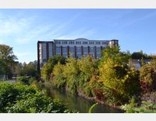 Ein mehrstöckiges Ziegelgebäude mit großen Fenstern im Hintergrund, davor ein schmaler Fluss, der von dicht bewachsenen Büschen und Bäumen in herbstlichen Farben gesäumt ist. Der Himmel ist klar und blau.