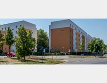 Mehrfamilienhäuser in einem Wohngebiet, bestehend aus mittelgroßen, rechteckigen Gebäuden mit orange-weißer und gelb-weißer Fassadengestaltung. Im Vordergrund befinden sich eine Verkehrsinsel mit Straßenschildern, Bäumen und parkenden Autos; eine ruhige Umgebung bei sonnigem Wetter.