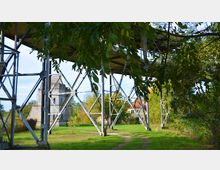 Metallkonstruktion in einer grünen Parklandschaft, teilweise von Bäumen umrahmt, mit einem steinernen Gebäude und einem Haus mit rotem Dach im Hintergrund. Ein schmaler, geschwungener Weg führt durch die Wiese unter der Konstruktion hindurch.