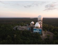 Ehemalige Abhörstation auf dem Teufelsberg in Berlin, umgeben von dichtem Wald. Die Szenerie zeigt mehrere weiße, kuppelartige Strukturen auf einem verlassenen Gebäude, mit Graffiti an den Fassaden und einer weiten Waldlandschaft im Hintergrund bei Sonnenuntergang.
