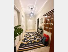 Flur in einer Wohnung mit hellen Wänden, einem geometrisch gemusterten Teppich und minimalistischer Einrichtung. An der rechten Wand hängen Wandhaken und ein Spiegel, daneben steht ein kleiner Glasregal mit Dekoration; eine Zimmerpflanze ist links sichtbar.