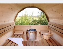 Innenansicht einer hölzernen Außensauna mit gewölbter Decke, Holzbänken und einem Saunaofen in der Mitte. Durch ein großes Fenster ist eine grüne Landschaft mit Bäumen zu sehen.