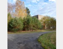 Eckiges Gebäude aus Beton mit kleinen Fenstern, teils von hohen Bäumen und Sträuchern umgeben. Im Vordergrund befindet sich ein asphaltierter Weg, der von einer Grünfläche gesäumt wird.