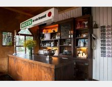 Gemütliche Bar mit rustikalem Holzthekenbereich, umgeben von Holzregalen, die mit Kerzen, Flaschen und dekorativen Gegenständen bestückt sind. Im Hintergrund ein Kreidetafel-Menü, ein Schild mit der Aufschrift „Holsten – der echte Biergenuss“ sowie Fenster mit Blick ins Grüne.