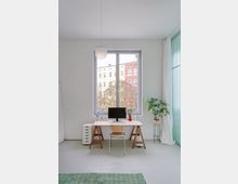 Helles, minimalistisches Homeoffice mit großem Fenster zur Straße, schlichtem Schreibtisch auf Holztresteln, Monitor und Lampe sowie einem einzelnen Stuhl. Rechts stehen eine hohe Zimmerpflanze und bodenlange weiße Vorhänge neben einer türkisfarbenen Glasfront.