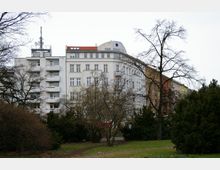 Mehrstöckige Wohngebäude mit weißen und farbigen Fassaden, teilweise mit Balkonen, hinter parkähnlicher Grünfläche mit Bäumen und Sträuchern. Der Himmel ist bewölkt, und Mobilfunkantennen sind auf einem der Gebäude sichtbar.