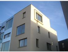 Ein modernes, mehrstöckiges Wohngebäude mit schlichtem, beigem Putz und großen Fenstern, steht in einer städtischen Umgebung unter klarem, blauem Himmel. Links daneben ist ein weiteres Gebäude mit einer silbernen Fassade und Glasfenstern sichtbar.