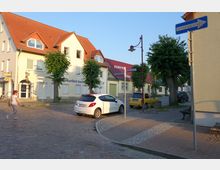 Straßenszene in einer kleinen Stadt mit Kopfsteinpflaster, zweistöckigen Wohn- und Geschäftshäusern mit roten Ziegeldächern und Bäumen am Straßenrand. Im Vordergrund sind parkende Autos, Straßenschilder und eine einzelne Fußgängerin zu sehen.