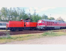 Zwei rote Diesellokomotiven stehen nebeneinander auf Gleisen in einem Bahnbereich, umgeben von grünen Bäumen und einem blauen Himmel. Im Hintergrund ist ein abgestellter weißer Zug zu sehen.
