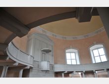 Innenraum einer Kirche mit hellen, pastellfarbenen Wänden. Der Raum zeigt eine umlaufende Empore mit weißen Säulen und Balustraden sowie eine weiße, schlichte Kanzel in einer halbrunden Wandnische unter einer Kuppeldecke.