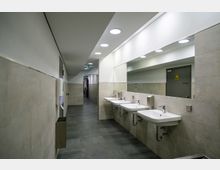 Ein moderner, hell beleuchteter Waschraum mit vier weißen Waschbecken, großen Wandspiegeln und beigefarbenen Fliesen. Im Hintergrund ist ein Korridor mit einer Tür zu sehen, an der ein Warnschild angebracht ist.